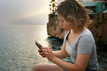 A young woman looks and writes on her phone against the background of a seascape. evening.High quality photo