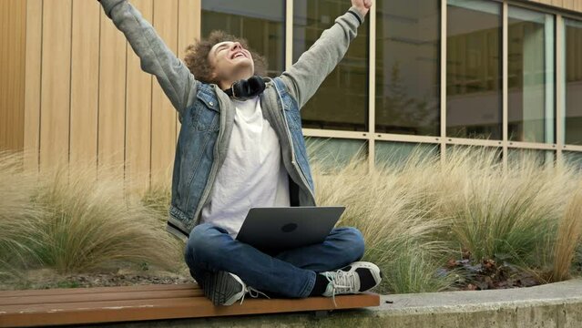 Guy sitting with a laptop on a bench in the yard of an educational institution is overjoyed at something. Winning a computer game, good news, good luck on the stock exchange.