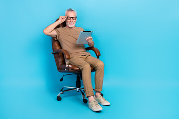 Full length portrait of cheerful aged man sit chair hand touch eyeglasses use tablet isolated on...
