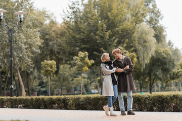 Fototapeta premium full length of redhead man and blonde woman in coat smiling while looking at each other in park.