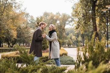 side view of joyful and young couple in coats holding hands while standing around plants in park. - Powered by Adobe