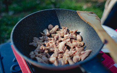 Chef is stir frying black roast pork on a picnic gas stove, camping menu, wooden ladle.