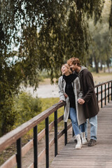 full length of redhead man and blonde woman in coat smiling while having date in park.