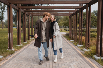 full length of happy young couple in autumnal coats holding hands while walking under multiple arch in park.