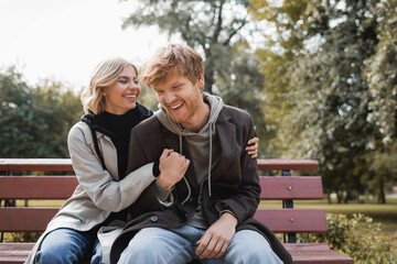 cheerful and blonde woman embracing redhead boyfriend while sitting on bench in park.