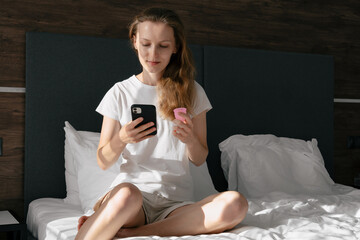 young woman sitting on bed and holding menstrual cup reading instructions on phone