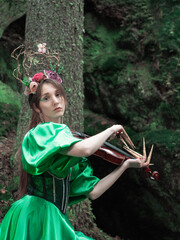 A beautiful woman in a long green dress with a corset and a decorative crown of branches and flowers on her head. The forest nymph plays the violin.