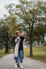 Fototapeta premium happy man in coat hugging cheerful young woman with closed eyes laughing in park.