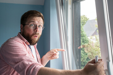 Young, attractive man stands at the window, looks at the camera in surprise and points finger into the yard. Close-up.