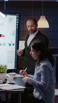 Vertical video: Workaholic businessman brainstorming marketing strategy overworking in company meeting office room late at night. Diverse multi-ethnic businesspeople looking at financial presentation