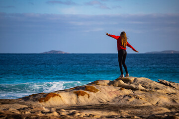 a beautiful long-haired girl stands on the rocks above the ocean with her hands raised enjoying the freedom; sunset on lucky bay beach in western australia
