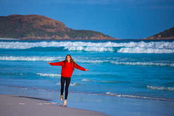 Fototapeta na wymiar A beautiful long-haired girl in a red sweatshirt walks on the sand at the famous lucky bay beach at sunset; a happy girl on the beach with white sand, turquoise water and hills in the background