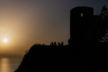 Grupo de amigos j&oacute;venes contemplando la puesta de sol y divirti&eacute;ndose junto a la Torre des Verger, un mirador junto al mar en las monta&ntilde;as de la Serra de Tramuntana de Mallorca.