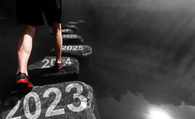 Young man walking crossing river on stones from 2023 to 2030 - obrazy, fototapety, plakaty