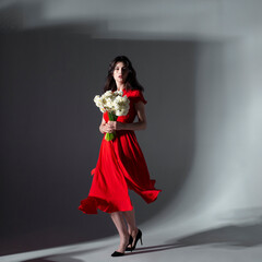 Full length image of young sexy woman posing in a red dress holding a bouquet of white daffodils, over grey background.