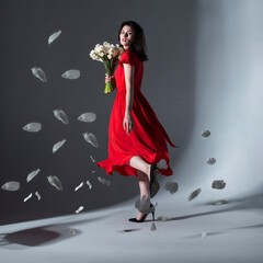 Full length image of young elegant woman posing in a red dress holding a bouquet of white daffodils, over grey background.