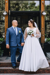 Fototapeta premium An adult stylish groom in a blue suit and a beautiful smiling bride in a white dress hold hands, against the backdrop of a city street in a park. Wedding photography of smiling newlyweds, portrait.