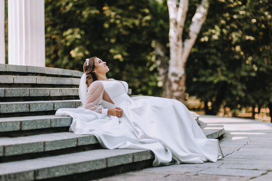 A beautiful, young bride in a white long dress is sitting on the steps in the park against the backdrop of ancient architecture. Wedding photography, portrait.
