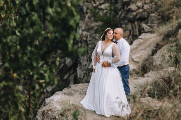 Fototapeta na wymiar A stylish groom and a beautiful young bride in a white dress are embracing in the open air, against the backdrop of rocks, mountain cliffs. Wedding photography, portrait.
