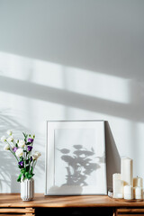 Modern minimalist Scandinavian style interior with white poster mockup, candles and flowers in vase on a wooden console under sunlight and home plants shadows on a white gray wall. Selective focus.