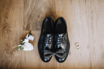 The groom's black shoes, bow tie, boutonniere, golden rings lie on a wooden background.Wedding photo of accessories and details, top view.