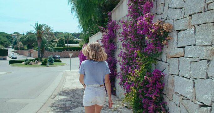 Camera follows pretty woman walking along a wall overgrown with blossom flowers. Cheerful and remarkable moment of summer vacation in Europe