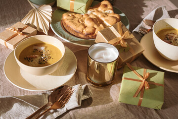 Festive table setting for a family dinner. Pumpkin soup, apple pie, candles, gifts on the table on a sunny autumn day.