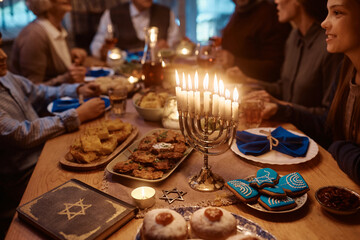 Lit candles in menorah on dining table with extended Jewish family in the background.