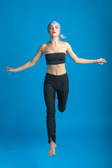 Fototapeta na wymiar Sport, fashion and make-up concept. Slim and beautiful woman with black and tight sport outfit, scarf and red lipstick jumping in air. Graceful pose freeze in motion. Blue studio background