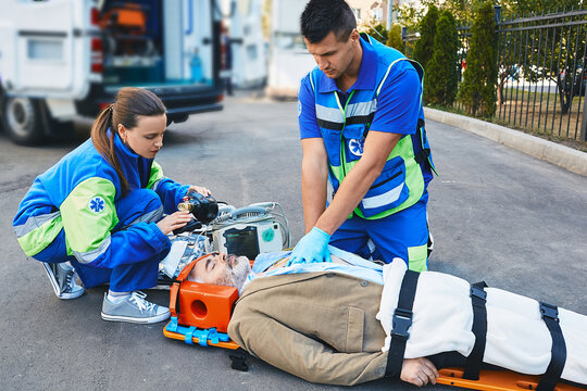 EMS paramedics performing closed-chest cardiac massage and artificial respiration for injured mature man who lies unconscious on medical stretcher near ambulance outdoor