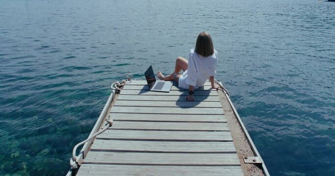 Woman sits relaxed on the pier next to laptop, looks at the sea, enjoys a summer calm day and remote work. Peaceful moment of happiness, satisfaction and freedom.