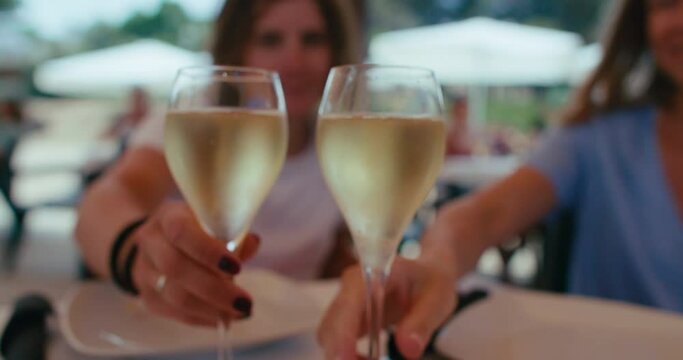 Closeup on glasses with cold sparkling wine at scorching summer day. Two girlfriends rise and cheer glasses, drink champagne, talk and smile together, while sitting on terrace of restaurant