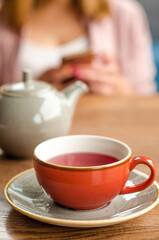 Red cup of red tea, a gray teapot stand on a table in a cafe. In the background, a young woman is working on her smartphone