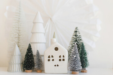Merry Christmas and Happy Holidays! Modern christmas scene, miniature cozy snowy village. Stylish little Christmas trees and houses decorations on white table. Winter banner, scandinavian decor