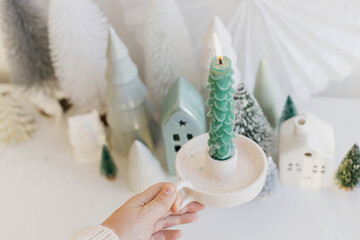 Merry Christmas! Hand holding stylish christmas tree candle on background of miniature winter village on white table. Modern little Christmas trees and houses. Holiday advent
