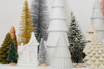 Merry Christmas! Stylish little Christmas trees and houses decorations on white table. Modern christmas scene, miniature cozy snowy village. Winter holiday banner, scandinavian decor