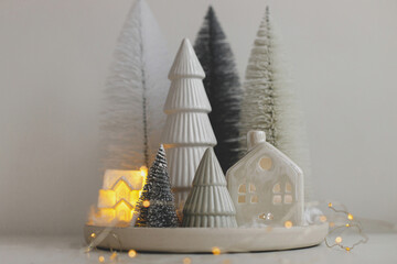 Merry Christmas! Stylish little Christmas trees and house on white table. Winter hygge, cozy christmas fairy scene, miniature snowy village with lights. Modern holiday decorations