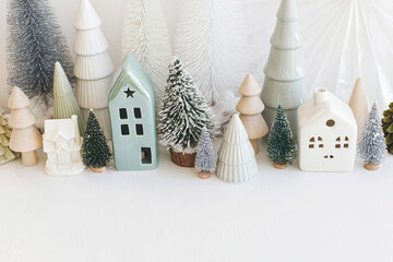Modern christmas scene, miniature cozy snowy village. Stylish little Christmas trees and houses decorations on white table. Merry Christmas and Happy Holidays! Winter banner, scandinavian decor - obrazy, fototapety, plakaty
