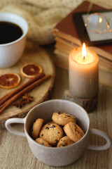 Bowl of cookies, cup of tea or coffee, chocolate, spices, knitted blanket, books, glasses and candle on the table. Cozy hygge atmosphere at home. Selective focus.