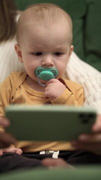One-year-old child sits in the arms of his mother and watches children's cartoons on the phone