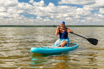 a woman in a closed swimsuit, a pareo and a headdress on her knees on a SUP board with a paddle floats.