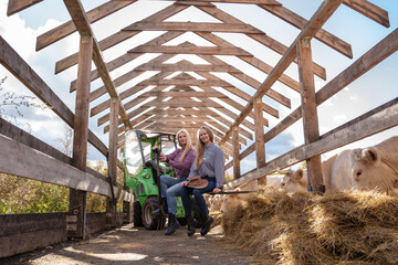 Portrait of two farmers women posing on farm vehicle around hay in barn.