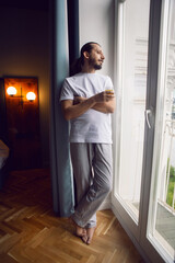 young man with a beard in a T-shirt and trousers barefoot stands with orange juice at the window in...