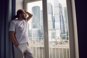 man with long hair, beard and glasses stands at a large window in an apartment with a view of a skyscraper in the morning. the guy is dressed in a white T-shirt and sweatpants barefoot.