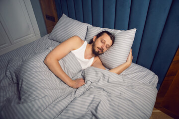 Obraz na płótnie Canvas Caucasian man with a beard and long hair in a white T-shirt is lying under a blanket and sleeping in a bed.