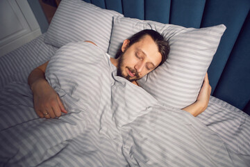 Caucasian man with a beard and long hair in a white T-shirt is lying under a blanket and sleeping in a bed.