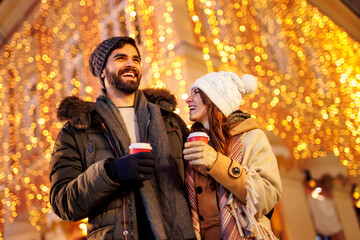 Couple drinking mulled wine and walking down decorated city streets while celebrating Christmas