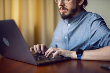 portrait of a businessman a man with a beard and glasses is sitting at a laptop at a wooden table at home. the freelancer works remotely next to the window the light is natural.