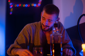 A young man with a beard and a tattoo is sitting in a bar. He has a glass of whiskey in his hands. Meetings with friends. Entertainment for adults. He smiles.