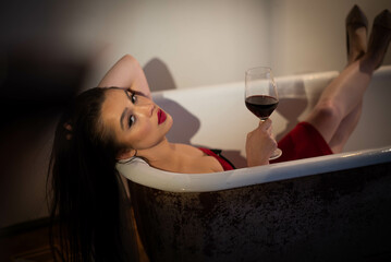 Fascinating brunette lies in the bathroom in a red dress. Looking at the camera with a glass of red wine.